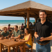 Imagen de un chiringuito en la playa con un ambiente veraniego, relajado y profesional. En primer plano, un camarero sonriente sostiene un PDA en la mano, simbolizando el uso de tecnología en la atención al cliente.